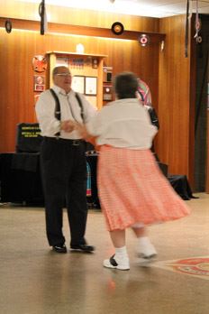 A man wearing suspenders dances with a woman in an orange skirt. 